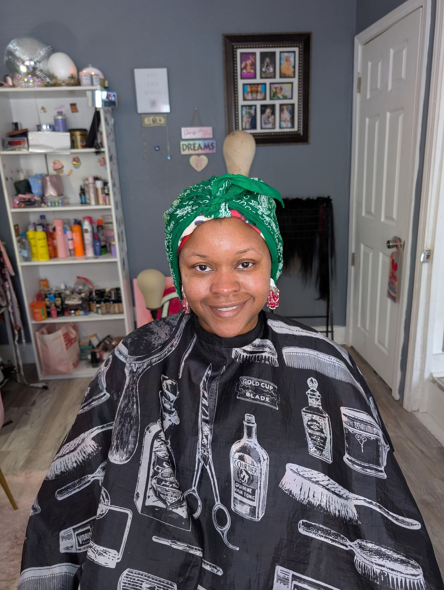 A smiling woman wearing a green headwrap and black barber cape sits in a hair salon.