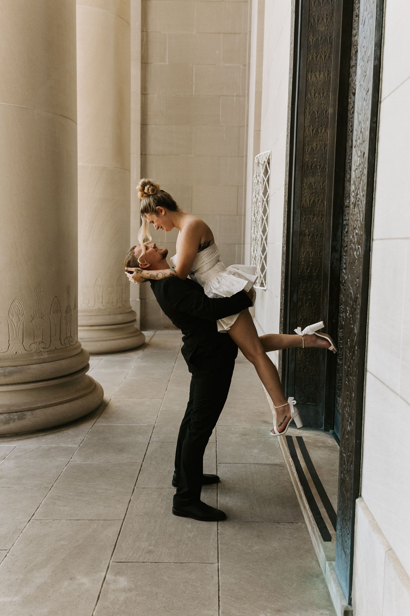 Groom lifting bride in white dress for a romantic kiss between stone columns at elegant venue.
