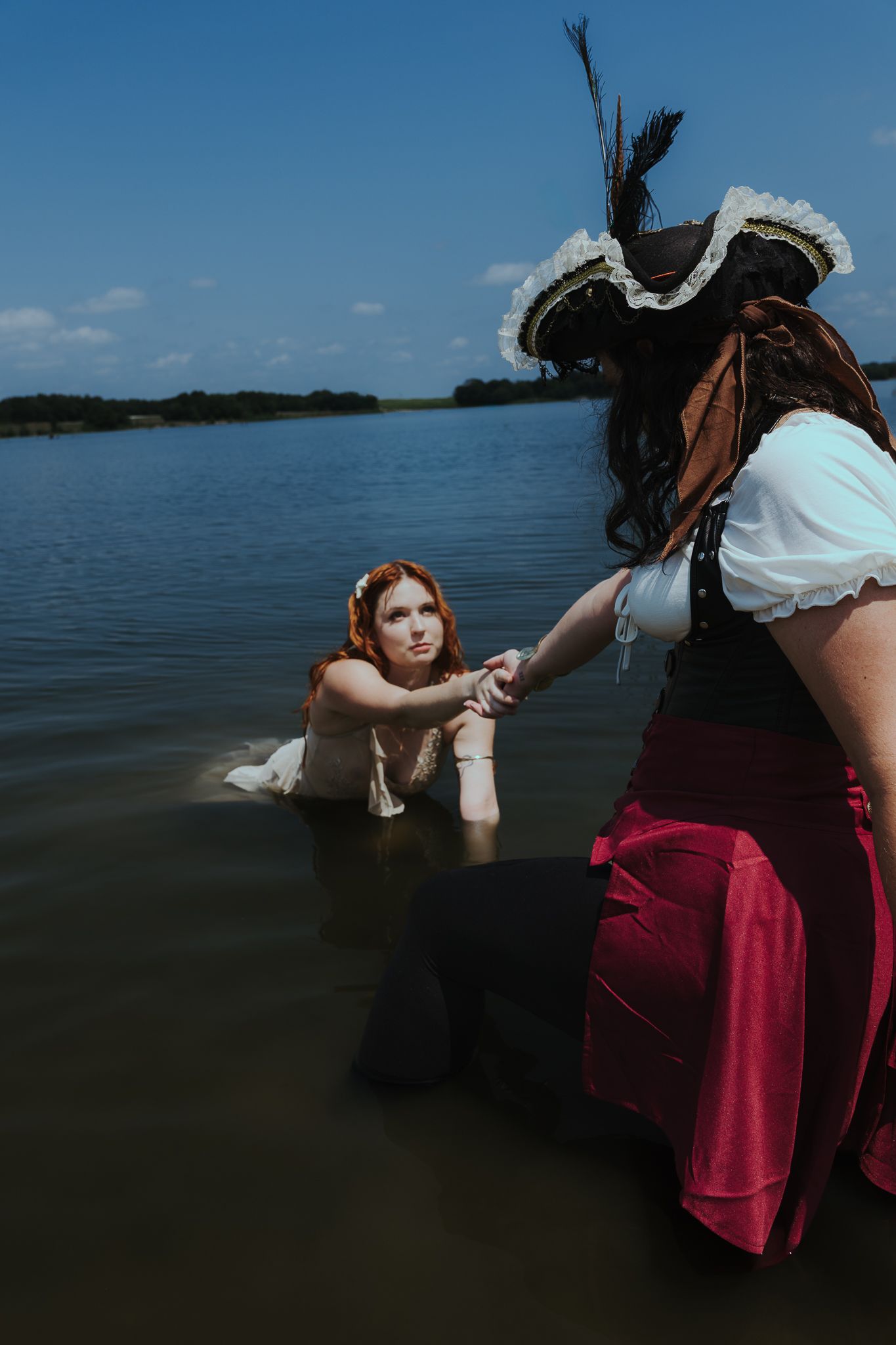 Two women in pirate costumes pose in a lake, one kneeling in water while the other reaches out her hand.