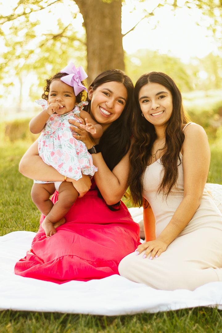 Family enjoys tender outdoor portrait session laying on white blanket in sunlit park setting with joyful expressions.