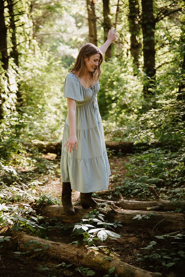 A figure in a light flowy dress explores a sunlit forest path among fallen logs and dappled woodland light.