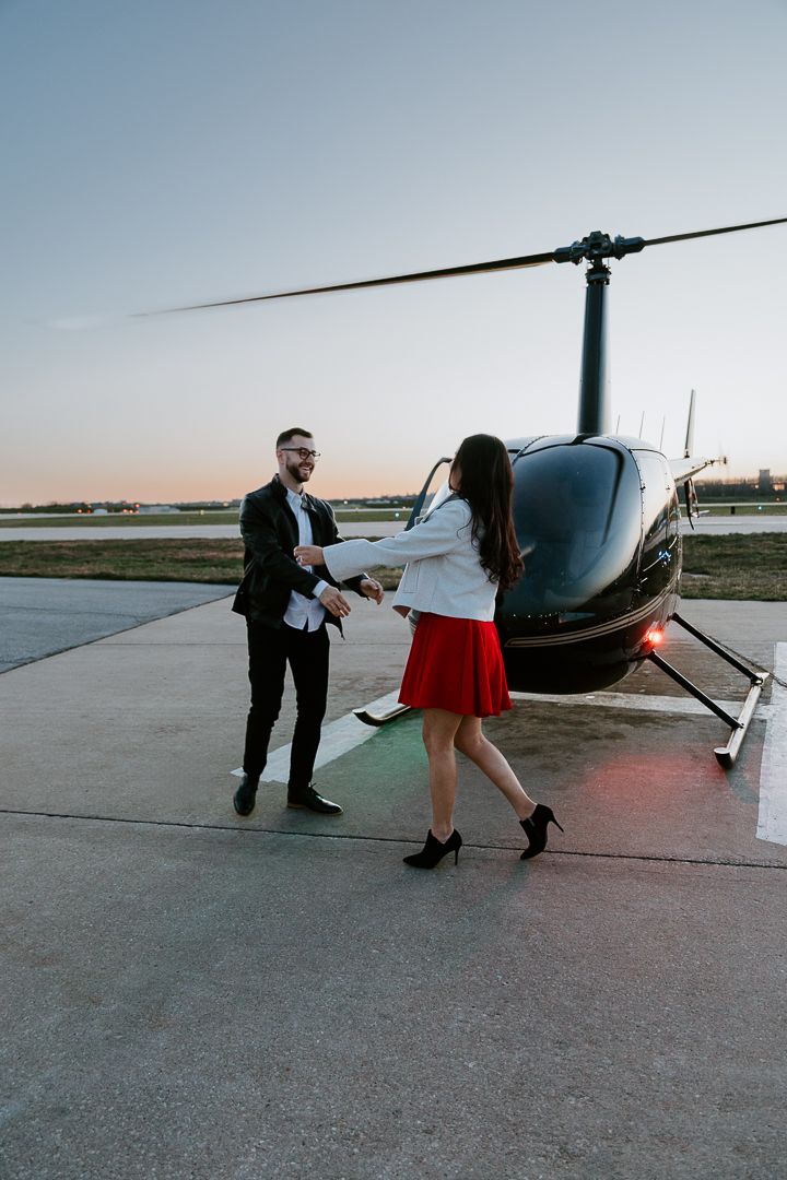 A couple stands next to a helicopter on a helipad during sunset.