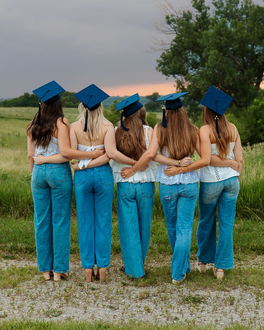 Group of graduates wearing matching blue caps and gowns pose with their backs turned against a grassy outdoor setting at sunset.