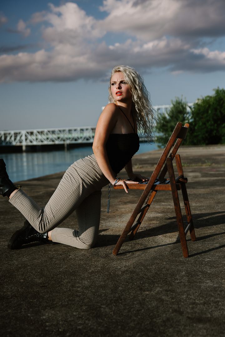 A fashion model poses with a wooden ladder near a bridge on a cloudy day by the waterfront.