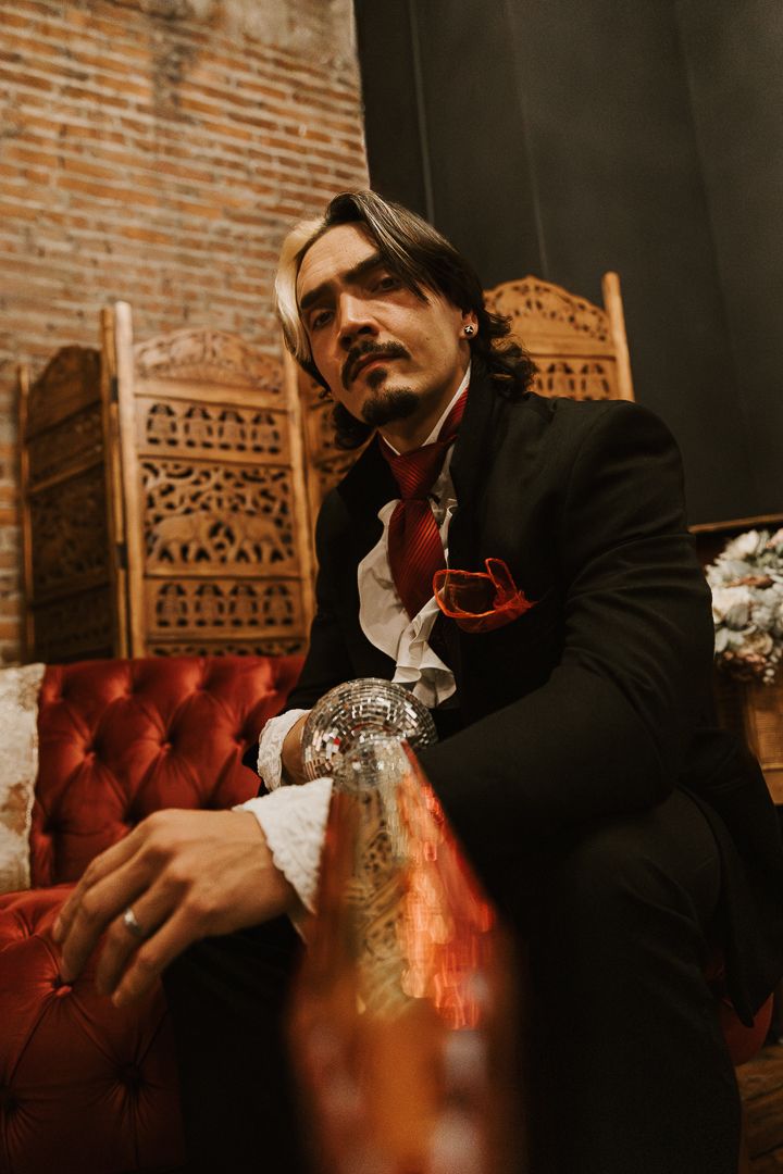 A person in dark attire relaxes on a red leather tufted sofa against exposed brick walls in moody lighting.