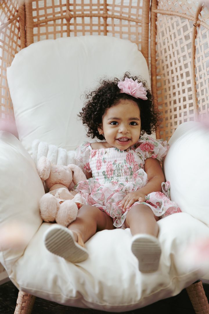 A child in a floral dress relaxes on a plush white chair with stuffed animals and pink accessories.