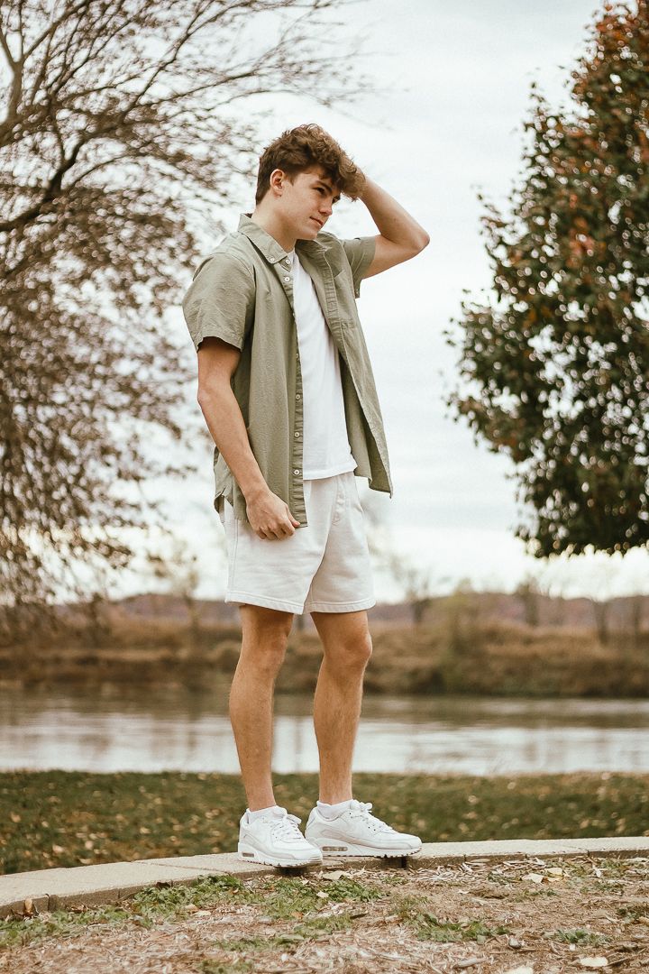 A casual outdoor photoshoot near a lake featuring white shorts and a beige shirt outfit in natural lighting.