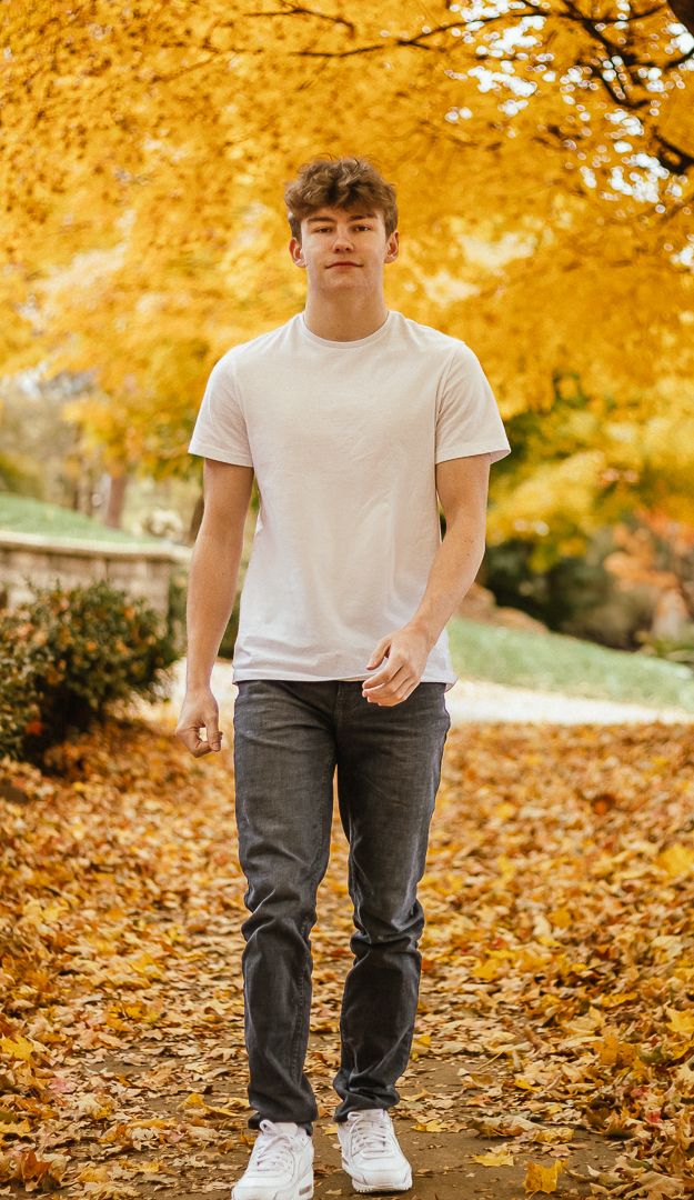 A person in a white t-shirt and gray pants walks through autumn leaves in a park with golden foliage above.