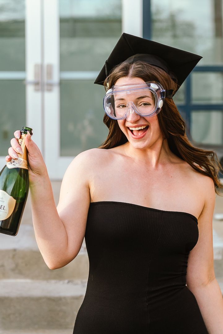 A graduate in a black dress and cap celebrates with champagne during a graduation photoshoot.