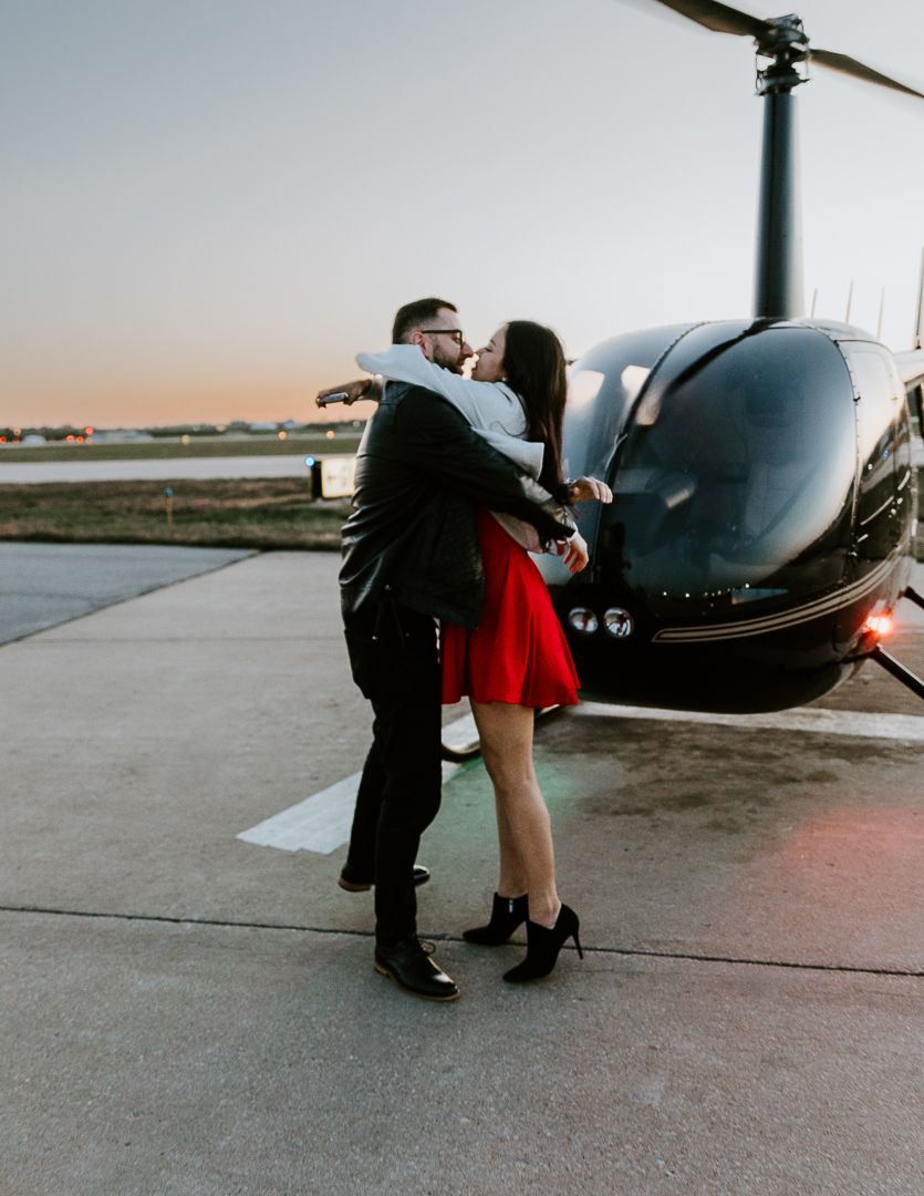 A romantic couple embraces next to a sleek helicopter at sunset on an airport tarmac.