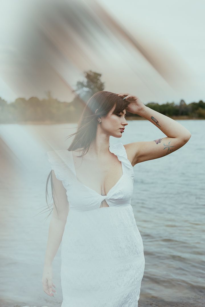 Ethereal figure in white dress poses gracefully by a serene lake during golden hour.
