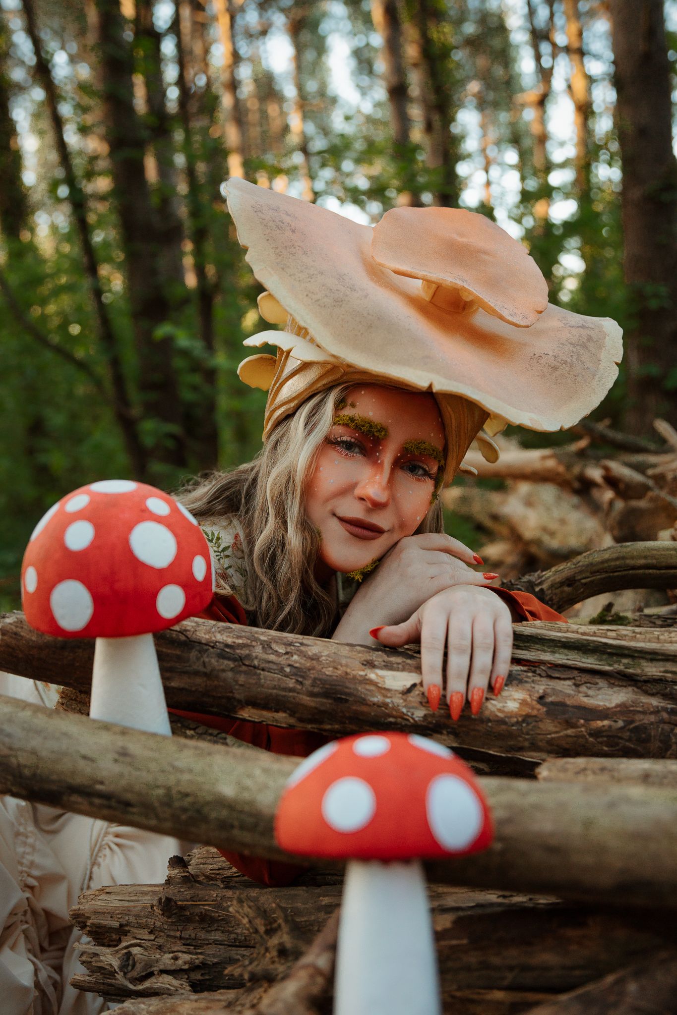 A woman in mushroom fairy costume with dramatic makeup poses among oversized toadstools in an enchanted forest setting.