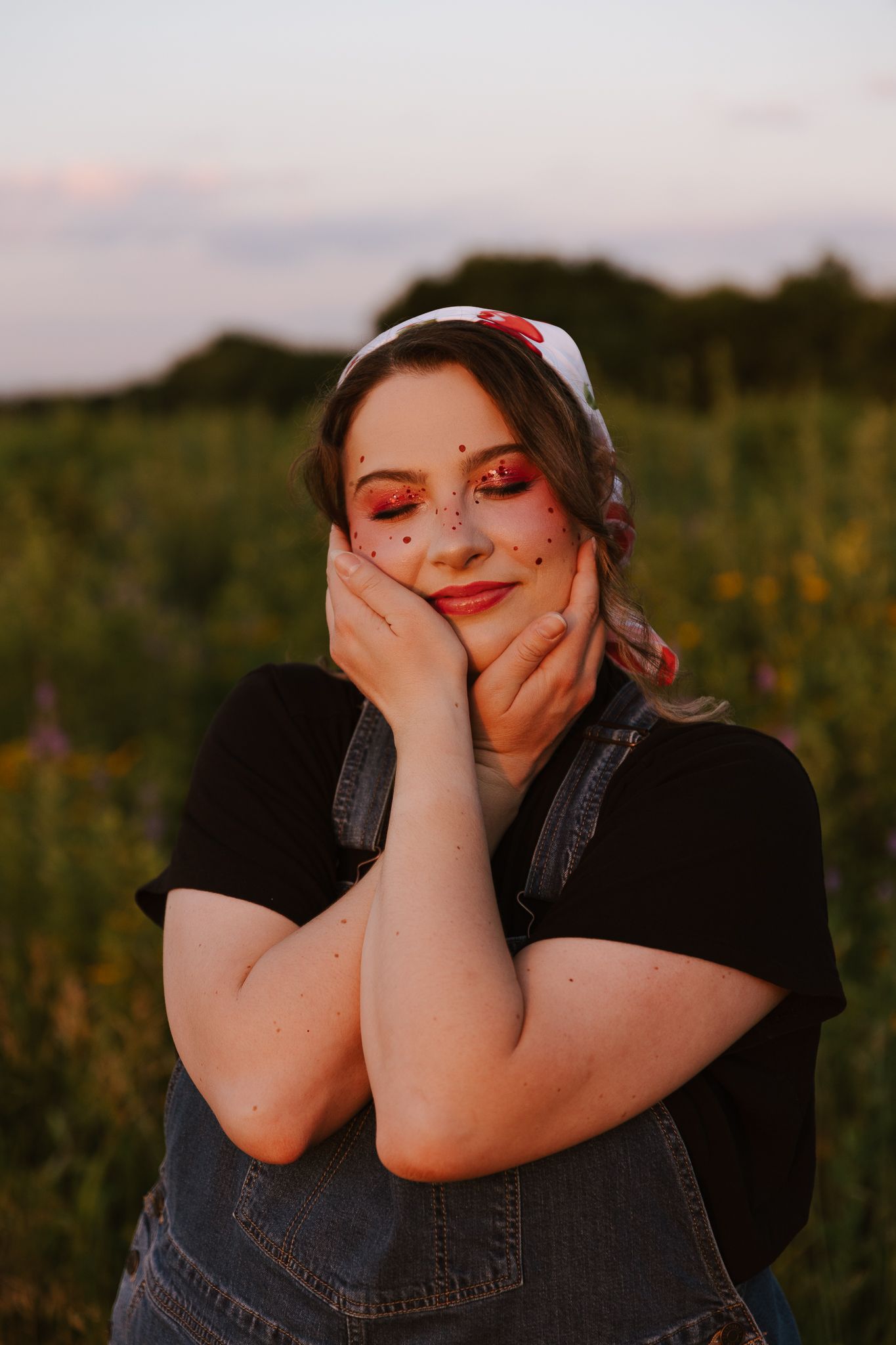 Young woman with cherry face gems and red eyeshadow poses dreamily in a sunlit field at golden hour.