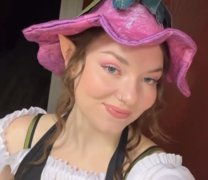 Person wearing pink renaissance-style hat with blue bow in selfie pose.