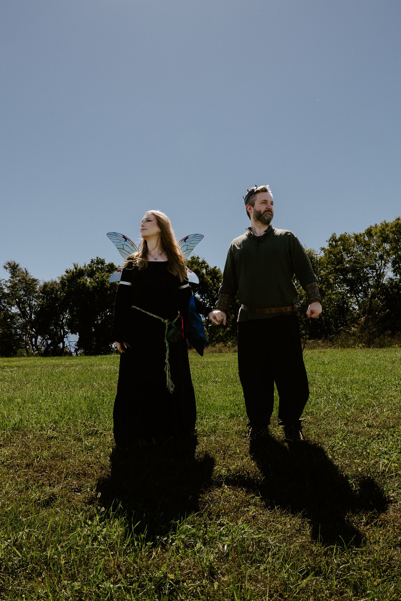 Couple in medieval-style costumes, one wearing translucent fairy wings, stand side by side on a sunny grassy field beneath a clear blue sky.