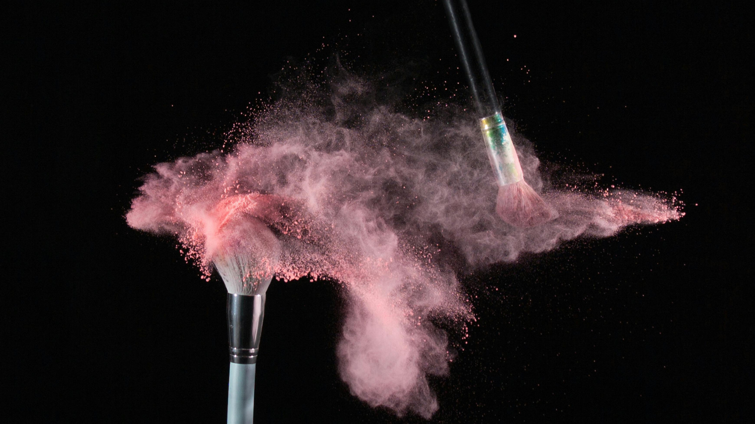 Two makeup brushes creating a pink powder explosion against a black background.