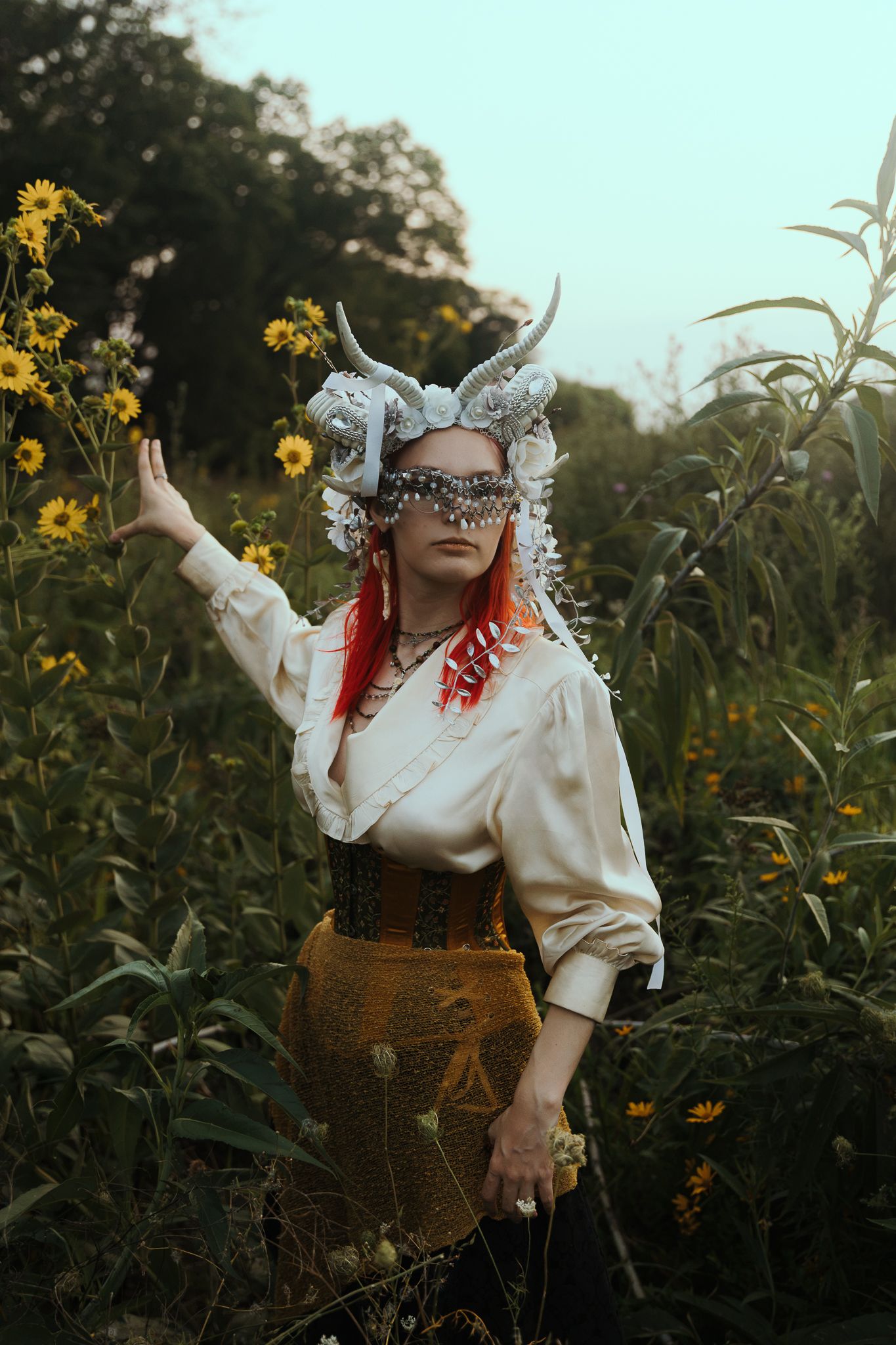 Woman in fantasy horned headdress and corset poses among yellow wildflowers in a mystical outdoor setting.