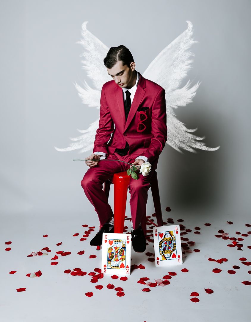 Figure in red suit with painted wings poses with playing cards and rose petals on light backdrop.
