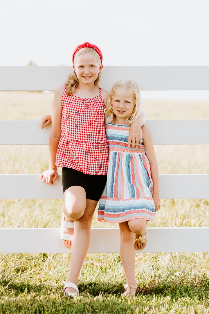 Two children in summer dresses sitting on white fence in golden field.