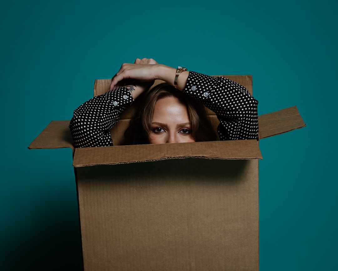 Person peeking through a cardboard box against a teal background wearing a polka dot outfit.