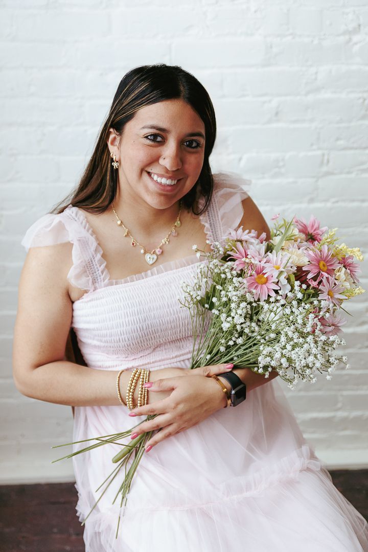 A woman in a pink dress sits against a white brick wall holding a delicate bouquet of baby's breath flowers.