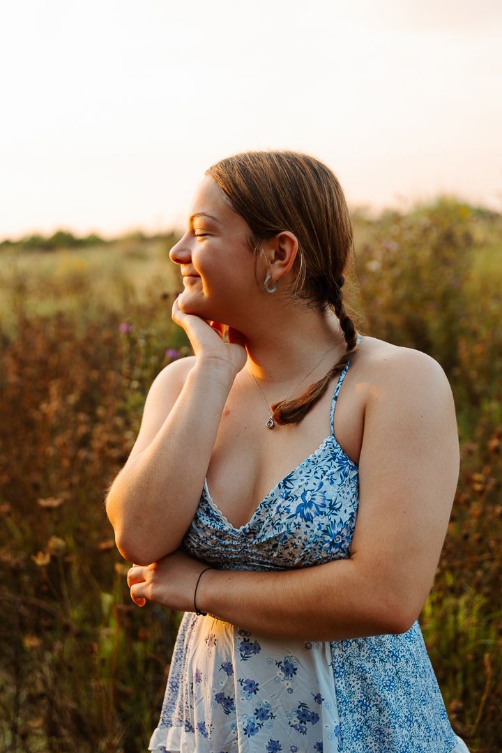 Person wearing a blue floral dress laughs joyfully in golden sunlight while standing in a natural outdoor setting.
