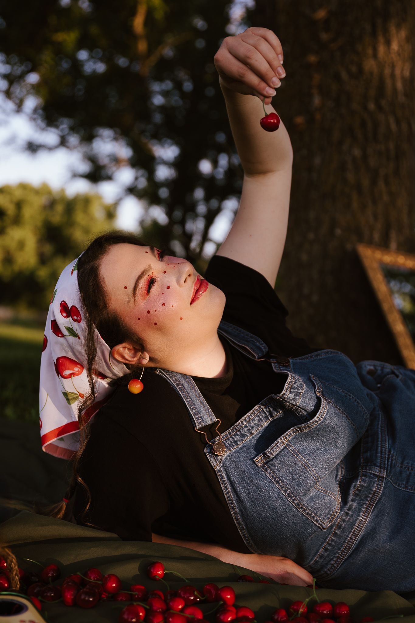 Young woman lying outdoors holding cherries above her face, wearing cherry bandana and denim overalls.