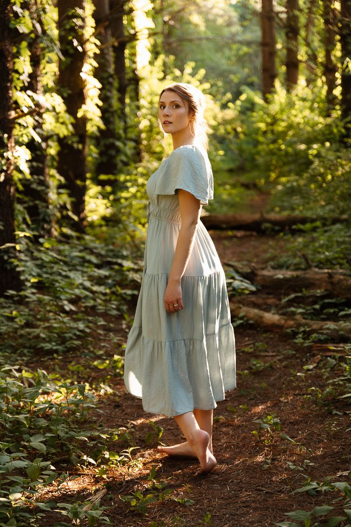 Person wearing a light blue flowing dress poses gracefully in a sunlit forest setting among trees and foliage.