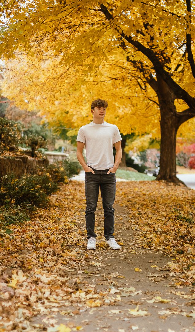 Person in white t-shirt and dark pants stands thoughtfully on leaf-covered path beneath golden autumn trees.