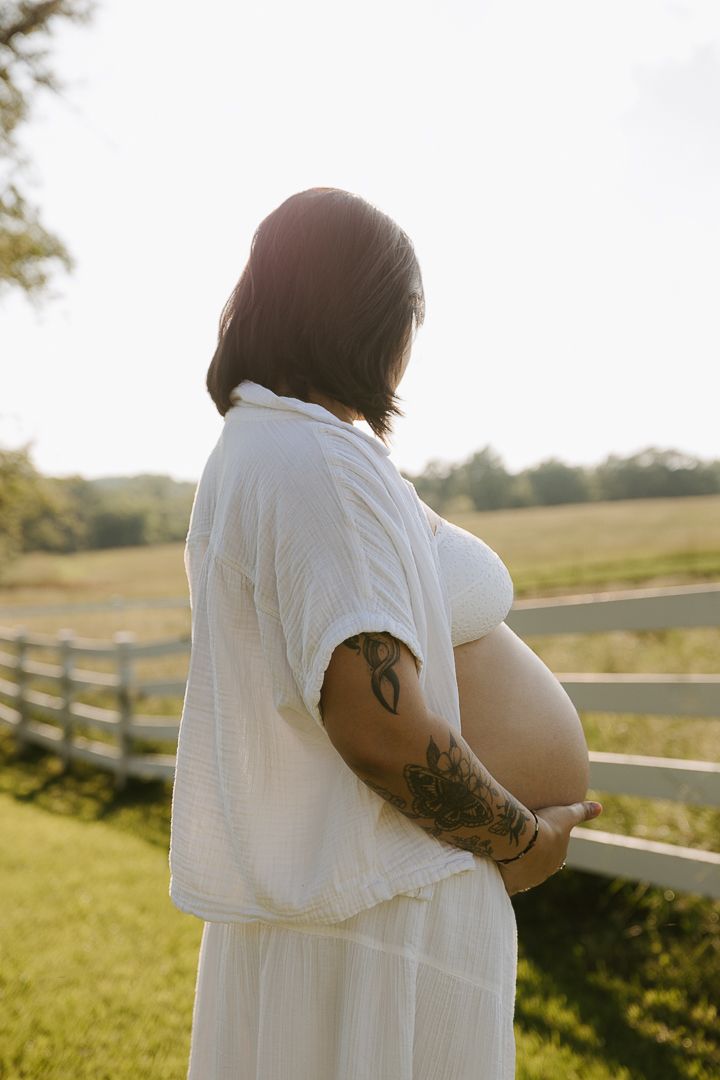 Back view maternity portraits showing tattoos and white dress with rustic fence and rolling hills in background.