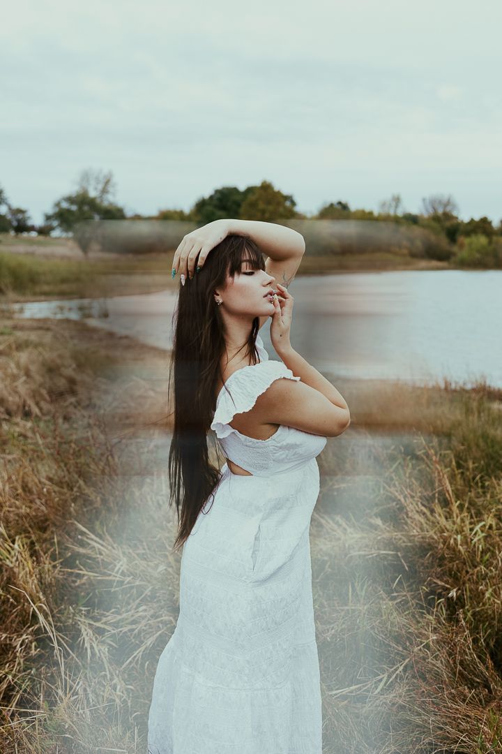 A dreamy photoshoot in a field by a lake features a flowing white dress and long dark hair blowing in the breeze.