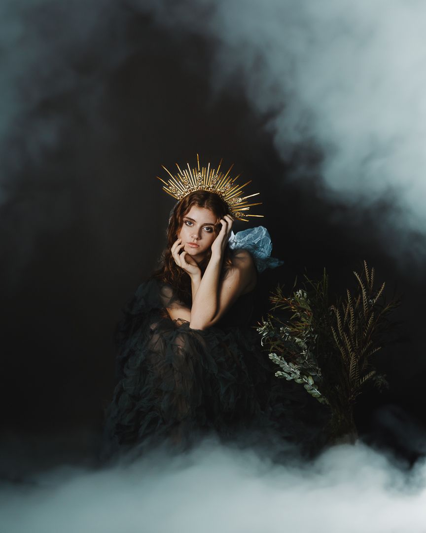 Woman in black lace gown and gold crown poses dramatically in misty studio with greenery.