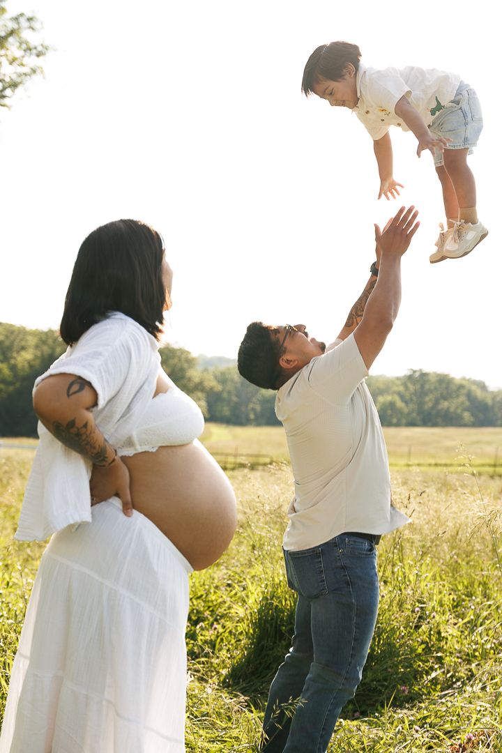 Family in a sunlit meadow with white maternity dress and casual attire playing together during golden hour.