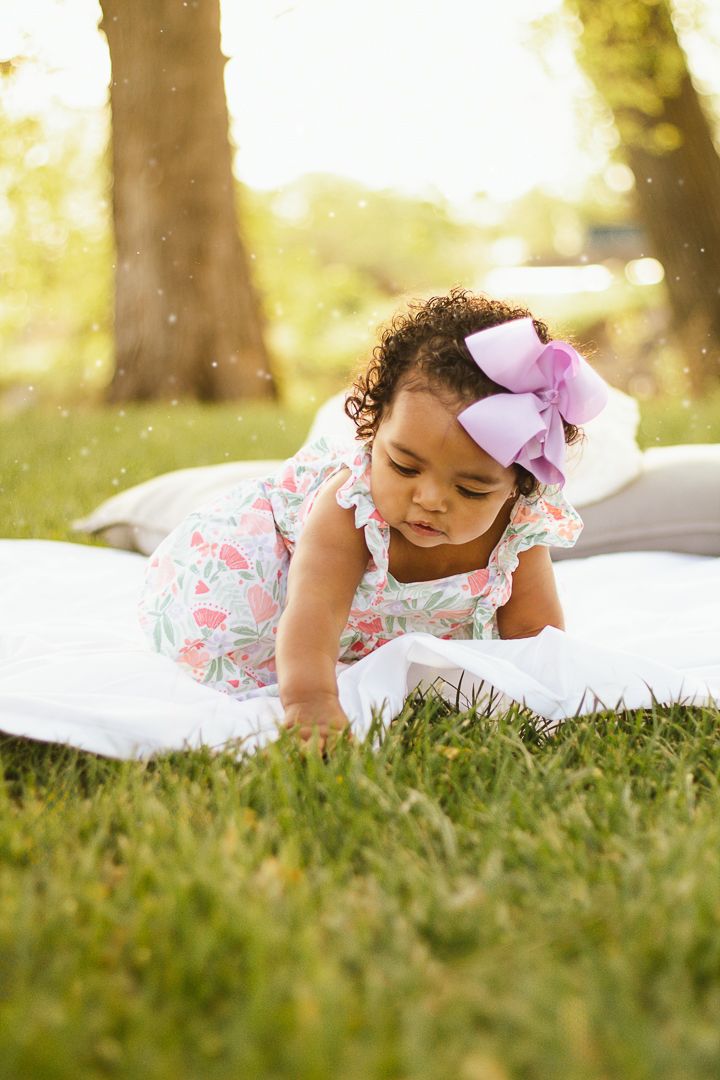 A small child in a floral dress lies on a white blanket on grass in a sun-dappled outdoor setting.
