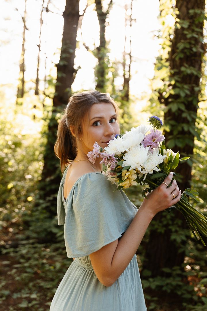 Bridesmaid in seafoam green dress smells bouquet of white and pink flowers in sunlit forest setting.