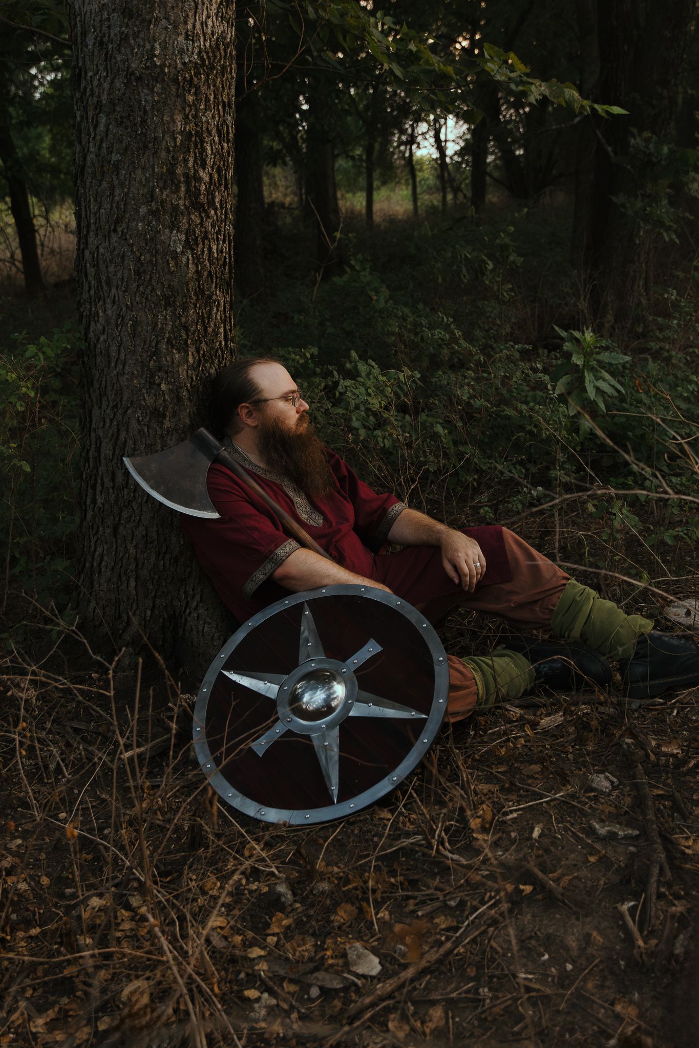 Bearded man in Viking costume resting against tree in forest with metal shield beside him.