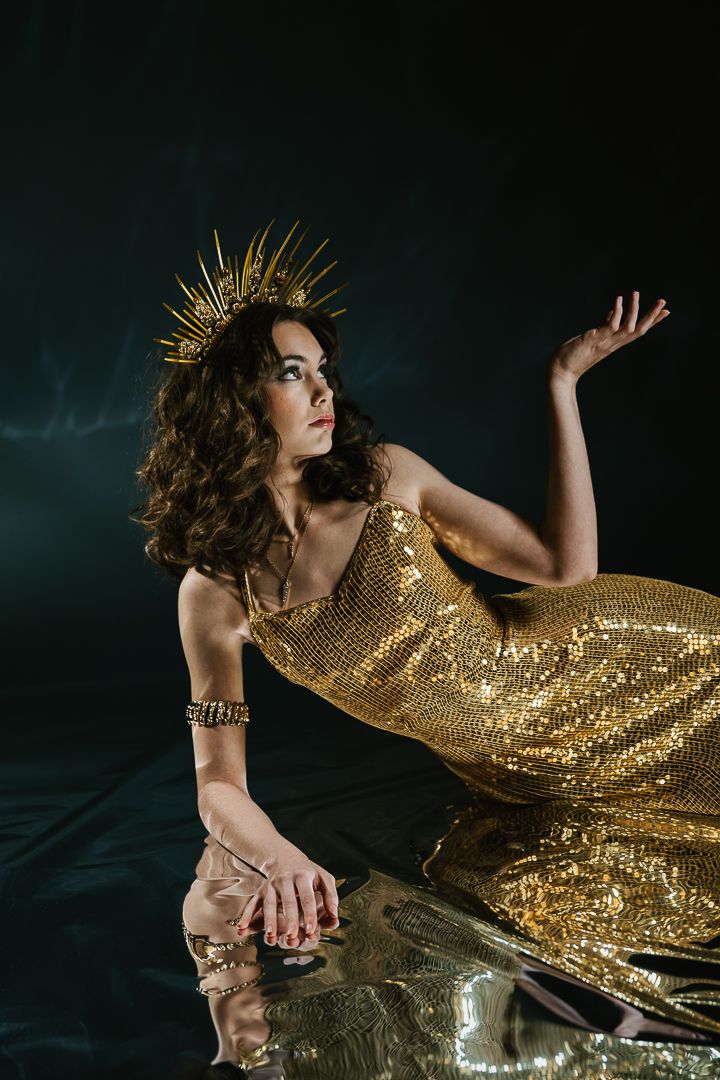 Dramatic fashion shoot with gold sequin dress and crown against dark background.