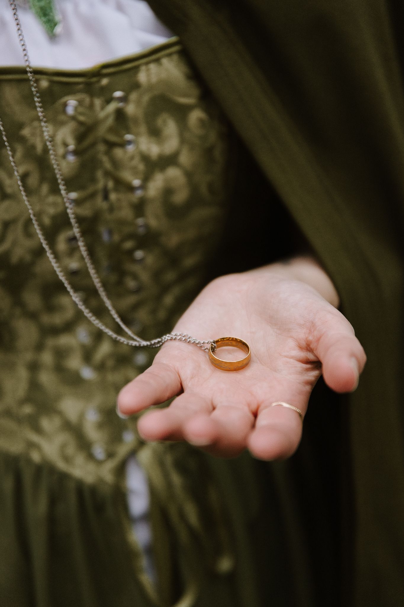 A hand holding a gold wedding ring on a chain, wearing a green embroidered corset dress.