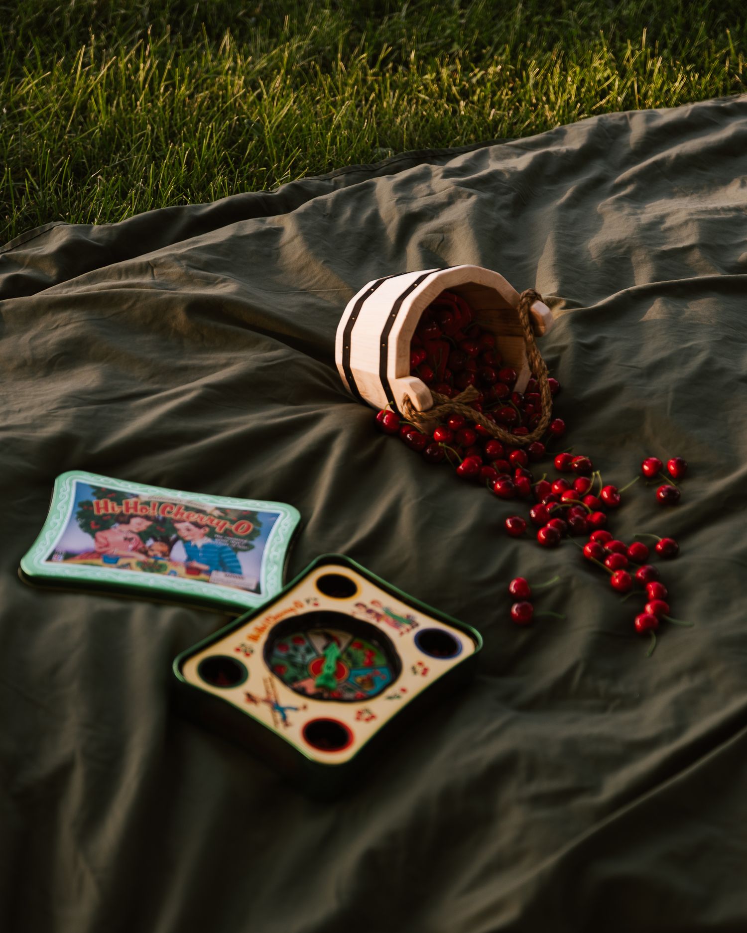 Spilled wooden bucket of red cherries on olive blanket with vintage board games outdoors.