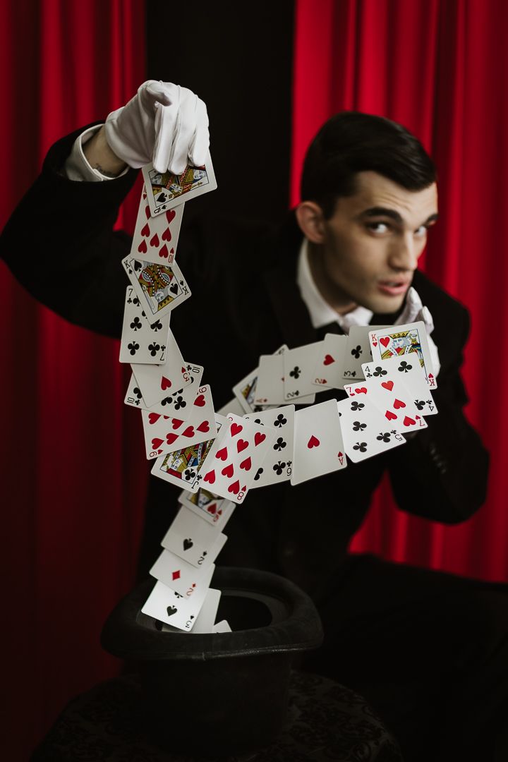 Magician in black suit and white gloves performing a card cascade trick with red curtain backdrop.