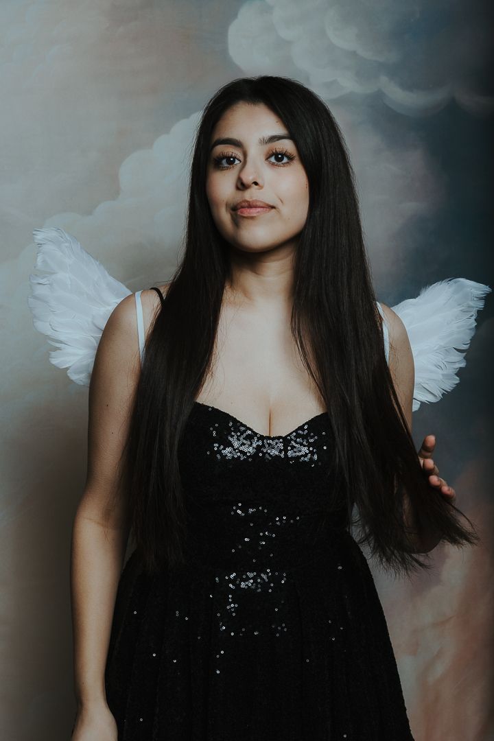 Woman with long dark hair wearing white angel wings and a black sequined dress poses against a painted cloudy sky backdrop.