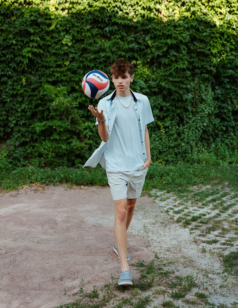 A person in a light blue shirt practices volleyball serves on a dirt court surrounded by green foliage.
