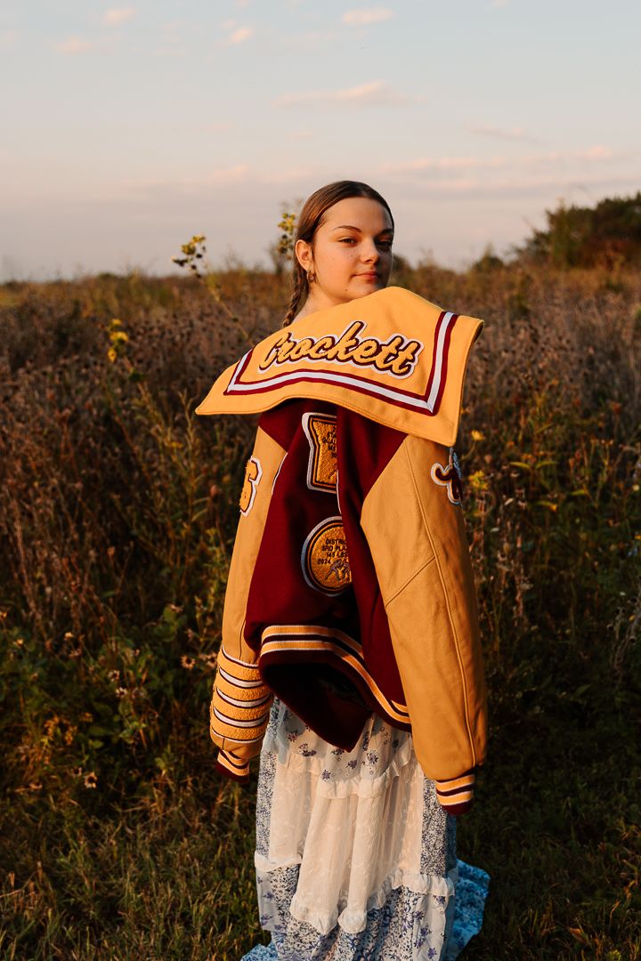 Someone wearing a vintage varsity jacket and blue floral dress poses against a dusky autumn field background.