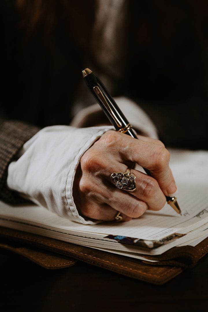 Close-up view of hands writing with a fountain pen on white paper against dark wooden surface in moody lighting.