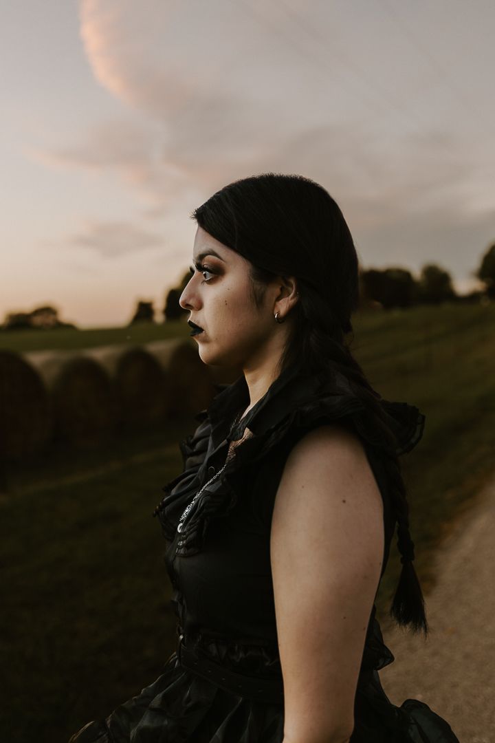 Dramatic profile portrait of someone in black clothing against a moody sunset landscape.