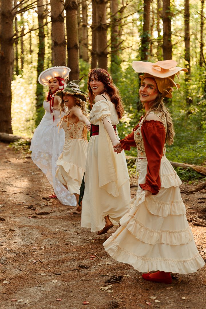 Three women in vintage Victorian dresses and decorative hats walk along a forest path in a whimsical outdoor photoshoot.