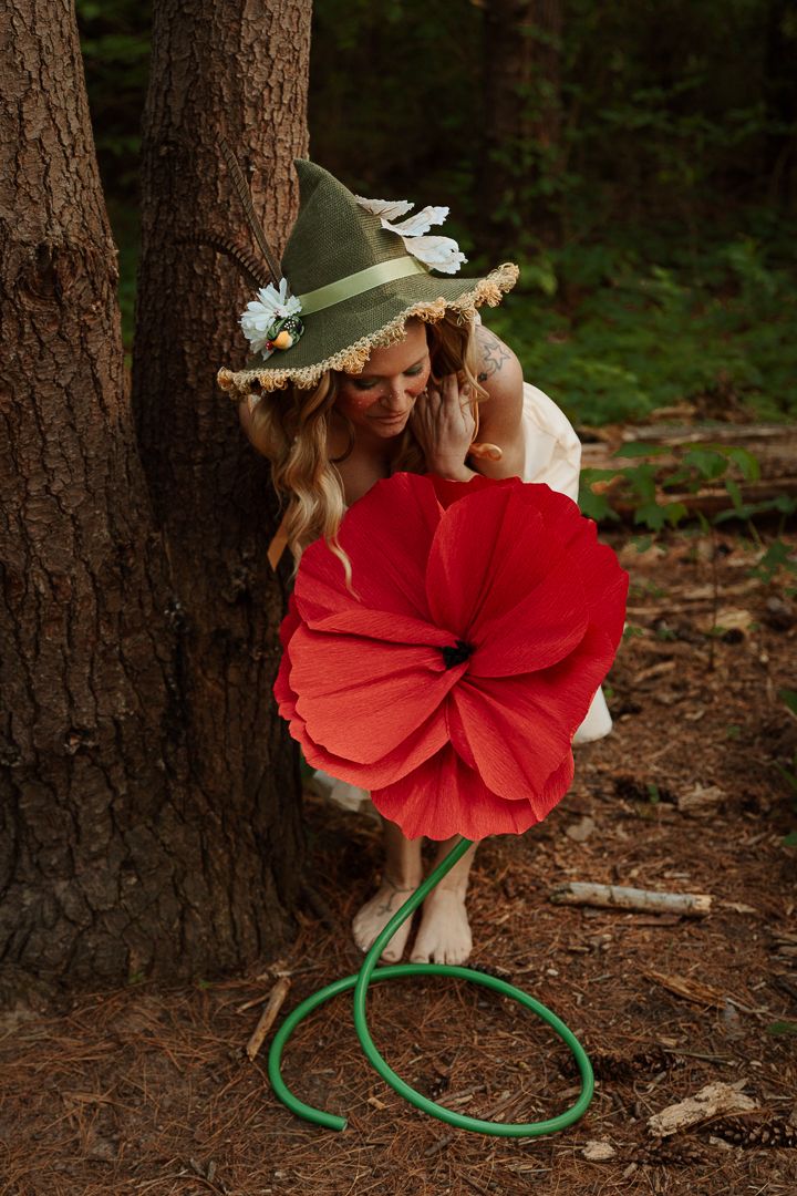 Barefoot woman in a green whimsical hat leans against a pine tree holding a giant red paper flower with a green hose stem in the forest.