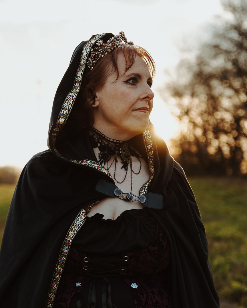 Woman in medieval black cloak and crown poses outdoors at golden hour sunset.