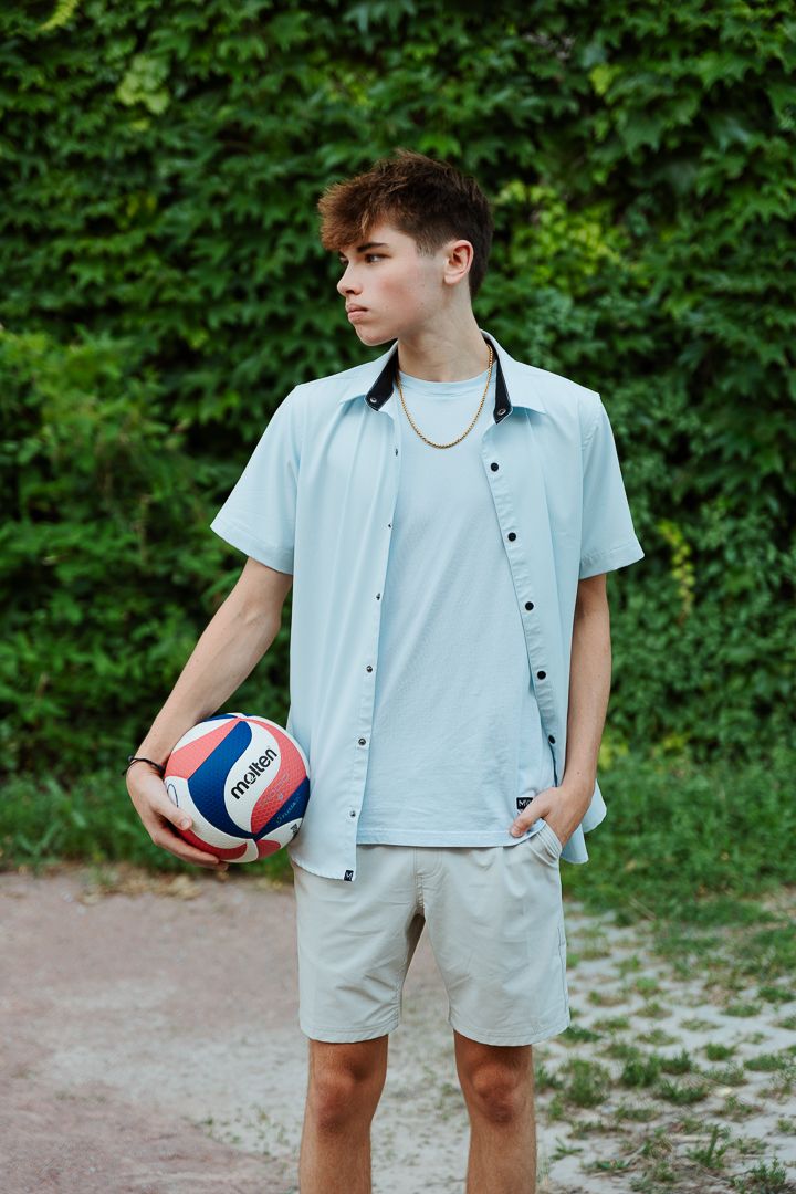 Person in light blue button-up shirt and beige shorts holds a volleyball in an outdoor setting with green foliage.