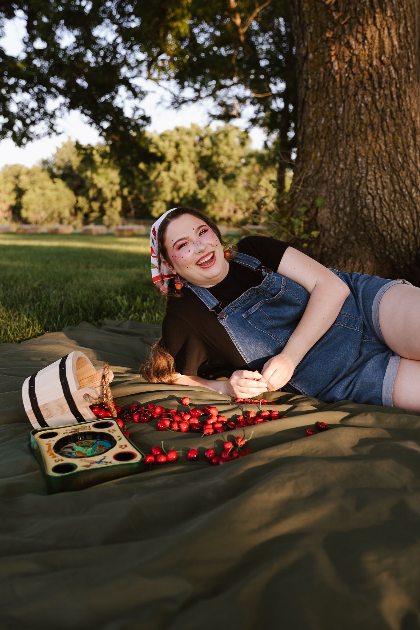 Young woman in denim overalls laughing while lying on a blanket with cherries under a tree outdoors.